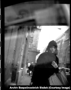 A young woman pulls her coat close as she passes a clandestine agent with a camera in downtown Prague.