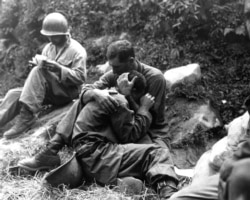 A grief-stricken U.S. soldier whose friend was killed in action is comforted by another infantryman in August 1950. In the background a corpsman fills out casualty tags.